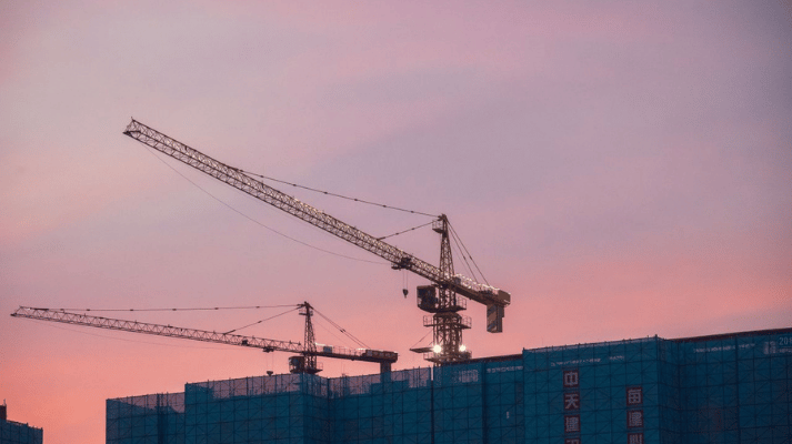 Construction cranes tower over a city skyline at sunset,
