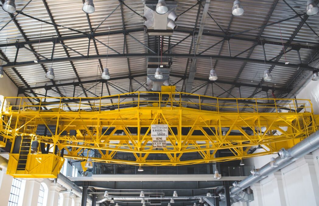A yellow gantry crane inside an industrial factory with metal roof and lights.