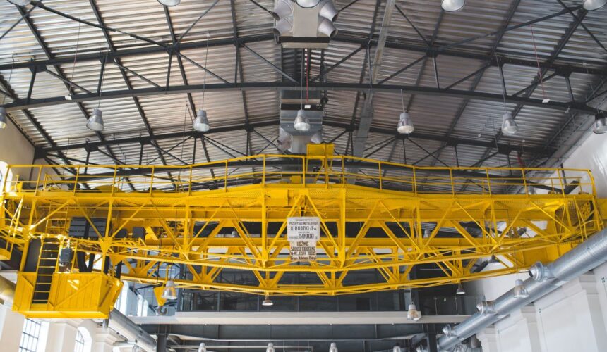 A yellow gantry crane inside an industrial factory with metal roof and lights.