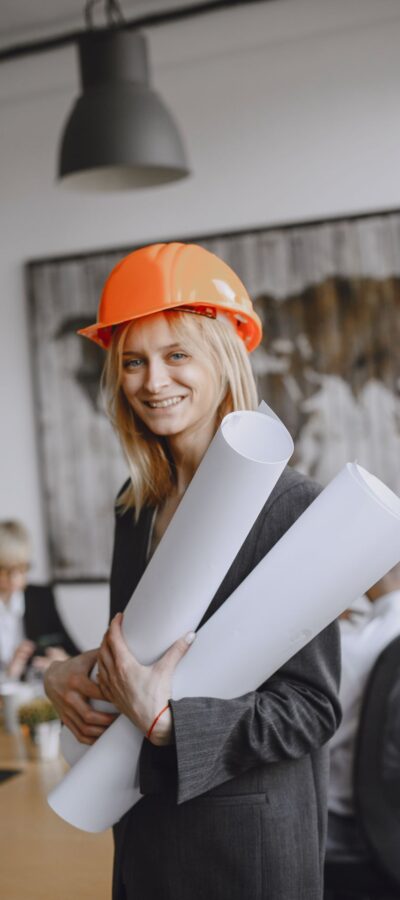 miling female architect in hard hat holding blueprints in modern office setting.