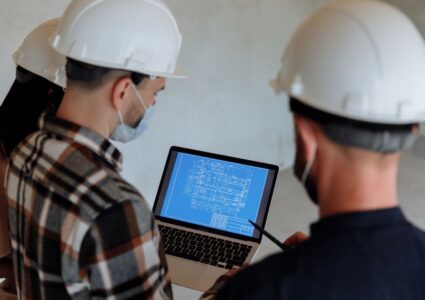 Engineers wearing hard hats and masks review a construction blueprint on a laptop closw